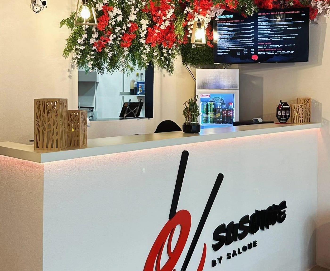 The Sushime counter, under a ceiling of red and white flowers, with the menu board above